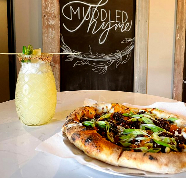 a craft cocktail in a pineapple-shaped glass sits next to a woodfired pizza in front of the Muddled Thyme sign